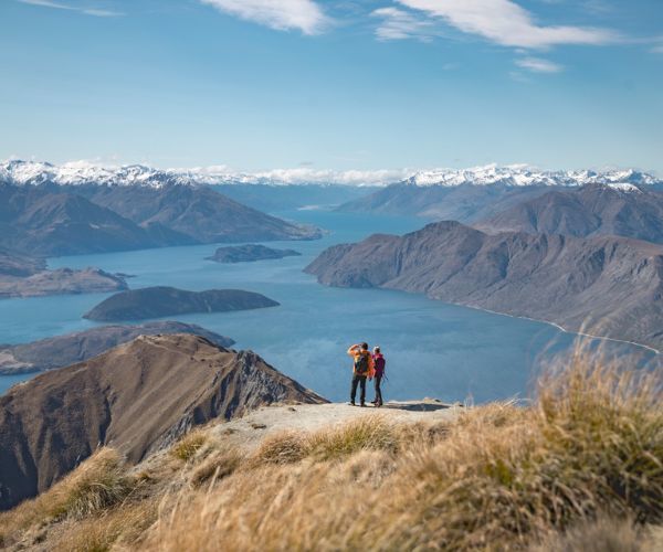 Two people standing on a mountain peak overlooking water and mountains