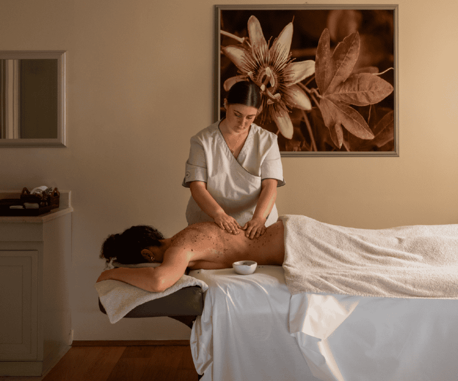 Calm spa treatment room with a therapist at Villa Biondi giving a full-body massage to a guest on a treatment bed, featuring soft lighting, neutral tones, and botanical artwork on the wall.