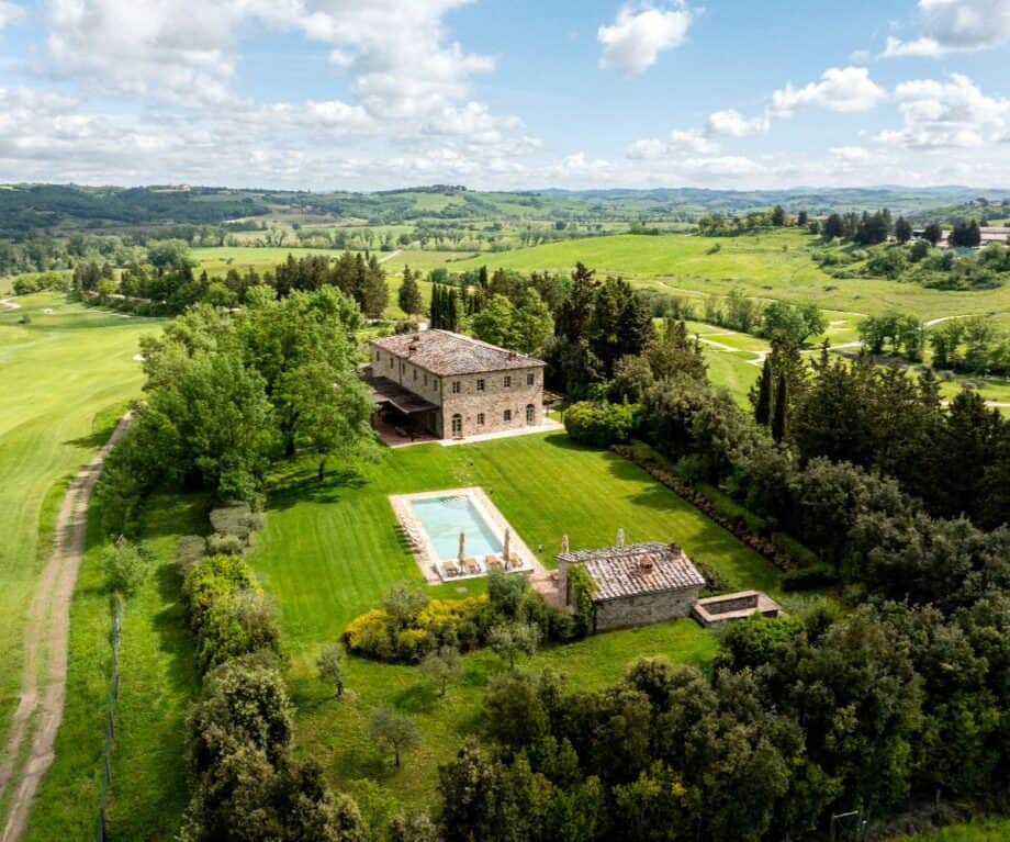 Aerial view of a traditional Villa Biondi in the Tuscan countryside, surrounded by rolling green hills, manicured gardens, tall trees, and a rectangular swimming pool with sun loungers.